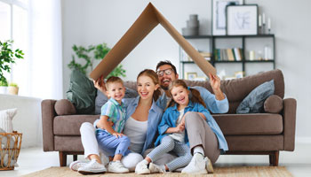A happy family sitting on teh floor of their living room with dad holding cardboard roof over their heads.